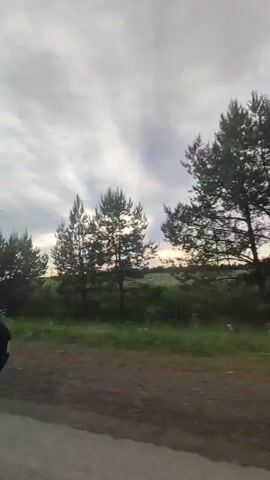 A car travels along a road next to a large green field under a cloudy sky at sunset. The sun is hidden behind the clouds, but its light shines through, creating an atmospheric effect. Trees line the edge of the field. A road sign is briefly visible. Music plays in the background.