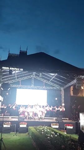 A video captures a nighttime outdoor performance on a stage with a large screen displaying text and graphics. A group of children dressed in traditional Romanian folk costumes are dancing and performing on stage. Behind them, musicians are playing instruments, suggesting a live orchestra. The stage is set up with lighting equipment and scaffolding. In front of the stage, there are decorative bushes and floral arrangements. The sky above is dark, indicating evening or night. The screen displays the text "CATALINA" and "Ansamblul Folcloric de Copii si Tineret Florile Moldovei Iasi."