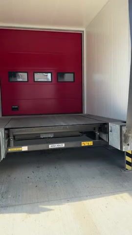 The video begins by showing the side of a loading dock, with a red overhead door and a metal platform extending outwards. The camera then pans to the side of a white semi-trailer truck with red and black stripes. A snowflake logo is visible on a white box attached to the underside of the trailer. The video continues to show a close-up of the trailer's rear door, which has a license plate that reads "R 1229 BDG". A hand then enters the frame holding a teal and black power tool, which is then used to tamper with the trailer's locking mechanism. The license plate reads "6864 MYI". The trailer appears to be a refrigerated unit, indicated by the snowflake logo and the overall design. The scene is outdoors, with a concrete ground and sunlight casting shadows. The overall context suggests a potential theft or tampering with a truck's cargo.
