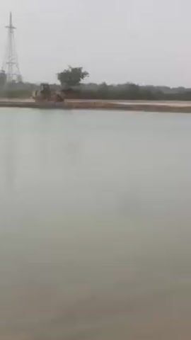 The video shows a body of water, likely a lake or river, with construction equipment on a sandy bank in the distance. There's a bulldozer, and a roller, and some cylindrical concrete structures. An electrical transmission tower is visible in the background. The sky is overcast, giving the scene a muted color palette. The water is calm and reflects the hazy sky. The scene appears to be a construction site near the water.
