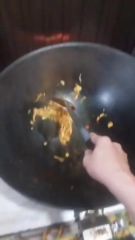 A person is using a spatula to stir-fry ingredients in a large, black wok. The ingredients appear to include shredded cabbage, tomatoes, and other small pieces of vegetables. The wok is placed on a stovetop, with a dark wooden wall visible in the background. The lighting is somewhat dim. The person's hand holding the spatula is visible, and the focus is mainly on the food being stirred within the wok.