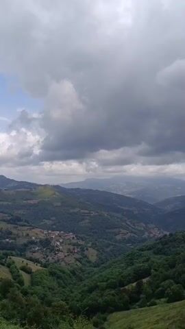 THE VIDEO SHOWS A LANDSCAPE OF ROLLING HILLS AND MOUNTAINS UNDER A CLOUDY SKY. GREEN VEGETATION COVERS THE SLOPES, WITH SMALL SETTLEMENTS VISIBLE IN THE VALLEYS. THE CAMERA APPEARS TO BE STATIONARY, OBSERVING THE VAST SCENERY.