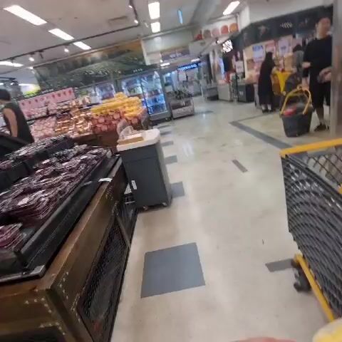 The video shows a person holding a yellow corn with a price tag in a grocery store. The corn is in plastic netting. The person is walking through the store with a shopping cart. The store is well-lit and stocked with various produce and other groceries. There are other shoppers in the background.