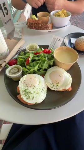The video shows a close-up view of two plates of food on a white table. The plate in the foreground contains two poached eggs on what appears to be smoked salmon and toast, served with a side salad of arugula and cherry tomatoes, a small cup of dressing, and a cup of creamy soup. The plate in the background has whole grain toast with avocado spread, a small bowl of avocado, and a bowl of scrambled eggs. A person's hand is visible in the upper right corner of the frame, holding a fork. Another plate with a bread roll is partially visible to the right of the foreground plate. The background suggests an outdoor seating area with brickwork visible. The lighting is natural and bright, indicating daytime.