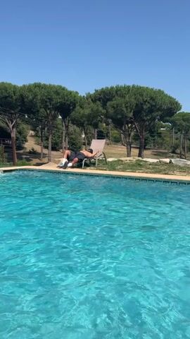 A person lies on a chaise lounge by a swimming pool. The person is wearing black shorts, white socks, and a black baseball cap. The chaise lounge is tan and is positioned at the edge of the pool. The swimming pool is filled with bright blue water. In the background, there are trees and a clear blue sky. The scene appears to be outdoors on a sunny day.