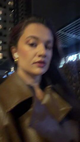 A young woman with long brown hair, wearing a brown leather jacket and gold hoop earrings, walks at night through a city. She has white earbuds in her ears and looks toward the camera as she walks. The background includes city lights, buildings, and storefronts.