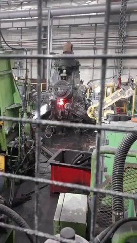 This media file captures an industrial setting, likely a factory floor, viewed through a wire mesh fence. The primary subject is a large, complex piece of machinery, possibly a forging press or a stamping machine, in operation. Sparks are visibly flying from the central part of this machine, indicating a high-energy process involving metal.

The machine itself is dark and appears heavily used, with various pipes, wires, and mechanical components. To the left and right of the main machine, other industrial equipment is visible, some painted green. A red bin sits in the foreground, containing what appear to be black hoses or pipes. The overall scene is dimly lit, suggesting an indoor industrial environment, possibly during a non-daylight hour, though the lighting is primarily artificial and functional.

The activity taking place is clearly an industrial manufacturing process, characterized by the intense sparks generated by the machinery. There are no people visible in the frame. The location context of Miryang-si, South Korea, implies this is an industrial facility within that region. No text is legible in the media file. The emotional tone is one of active, potentially hazardous industrial work, conveyed by the visual intensity of the sparks and the heavy machinery.