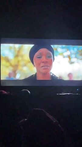 The video shows a television screen displaying a medium shot of a dark-skinned woman with tears in her eyes. She is wearing a black headscarf and dark clothing. Behind her, the background is blurred but appears to be outdoors, with trees and other people visible. The rest of the scene is dark, with only a partially visible figure in the foreground and the TV itself illuminated.