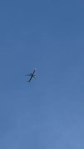 A wide shot captures a commercial airplane soaring through a clear, bright blue sky. The aircraft is a white passenger jet with dark-colored engines visible beneath its wings. It is angled slightly, with its nose pointing upwards and to the left of the frame, suggesting it might be ascending or banking. The plane's tail fin is also visible, showing a dark stripe near the top. The sky is uniformly blue with no clouds, indicating fair weather. The image is taken from below, looking up at the plane, which appears to be at a considerable altitude. There are no visible people, buildings, or ground details, making the plane and the sky the sole subjects. The time of day appears to be daytime, likely midday or afternoon, given the brightness of the sky and the clarity of the light. There is no visible text in the frame. The overall impression is one of peaceful flight against a serene backdrop.
