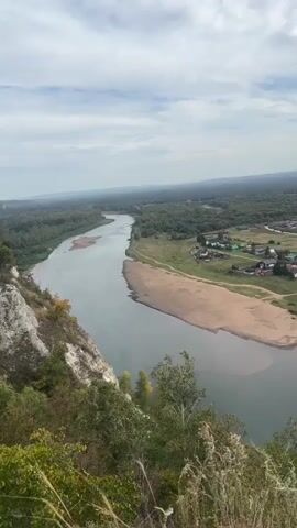 A high-angle panning shot captures a wide, calm river flowing through a valley. The river has a sandy island in the middle and is surrounded by dense green forests. On the right bank, a small village with houses and fences is visible. The sky is partly cloudy with a mix of white clouds and blue sky. The foreground shows the edge of a rocky cliff with some dry grass and foliage. The overall scene suggests a rural, natural landscape.