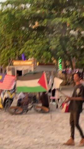 The video shows a beach scene with two flags, the Malaysian flag and the Palestinian flag, prominently displayed. A yellow scooter is parked near the flags. Several people are visible in the background, some sitting under a makeshift shelter and others standing. There are also tables set up with goods for sale, suggesting a market or vendor area. Trees and buildings are visible in the background, indicating an outdoor setting possibly near a resort or town. The overall impression is of a lively beach environment with commercial activity.