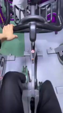 From a first-person perspective, the video shows a man on an elliptical machine in a gym. The man is wearing black leggings and has his feet secured on the pedals. The elliptical machine is gray with purple accents. The camera angle changes to show the man from the side as he works out on the machine. He is wearing white pants and a black hoodie. Other elliptical machines are lined up next to his. There are bodybuilding posters on the wall behind him. The gym appears to be well-equipped with various fitness machines.