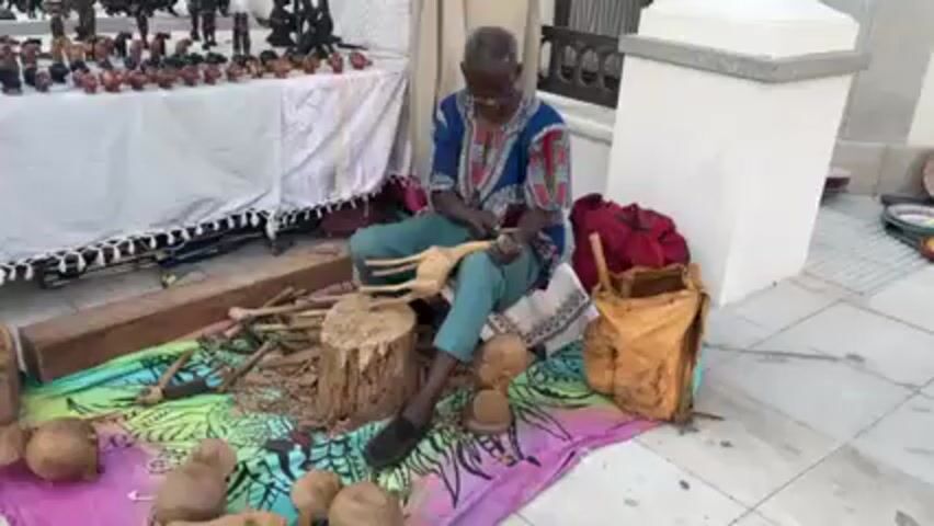 An elderly Black man is sitting cross-legged on a colorful blanket, carving a wooden animal figure. He is wearing glasses, a patterned blue shirt, and green pants. Around him are various wood carving tools, rough logs, and partially carved wooden figures. Behind him, a table displays finished wooden carvings. A bag is to his right, and a white pillar stands nearby. The scene is outdoors, on a paved surface.