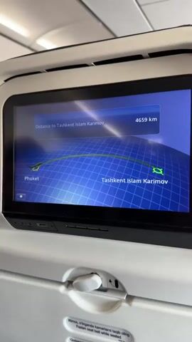 The video displays an airplane seatback screen showing a flight map. The screen indicates "Distance to Tashkent Islam Karimov" and "4659 km." A digital map with a blue grid shows a flight path, marked by an airplane icon at "Phuket" and a destination icon at "Tashkent Islam Karimov." Below the screen, part of a white seat-mounted tray latch is visible, along with a sticker with text in an unfamiliar alphabet.