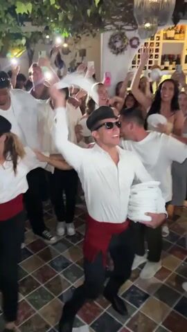 The video shows a crowded indoor gathering, possibly a party or celebration. A man in the foreground, wearing a white shirt, black pants, sunglasses, and a red sash, is enthusiastically waving a white cloth in the air while holding a stack of white items in his other arm. He appears to be dancing or leading a dance. Around him, other people are visible, many holding up their phones and appearing to film or photograph the event. Some are dancing, smiling, and cheering. The environment includes decorative greenery hanging from above and a tiled floor. The overall atmosphere is lively and celebratory.