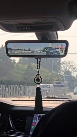 The video is filmed from the interior of a moving car, looking forward through the windshield and at the rear-view mirror. The rear-view mirror shows traffic on a road with several cars visible. A decorative hanging charm with beads and a tassel is suspended from the mirror. Outside the car, trees and buildings are visible. The dashboard of the car is also visible, including a screen displaying what appears to be a navigation interface. The overall lighting suggests it is daytime, possibly with some haze in the air.