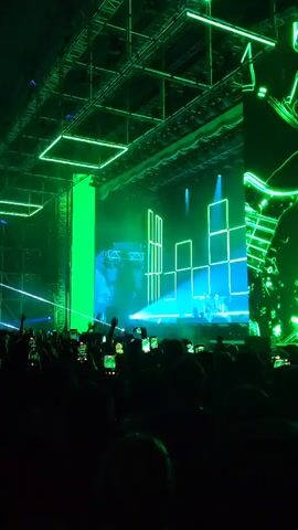 The video shows a concert stage illuminated with green and blue lights, with a large crowd of people holding up their phones to record the performance. The stage features a backdrop of square shapes and a vertical green light panel. Laser beams shoot across the space above the crowd. The audience is mostly silhouetted against the bright stage lighting.