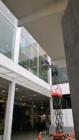 Two workers are on top of a bright orange scissor lift while cleaning the tall glass windows of a multi-story building. Traffic cones surround the base of the lift on the lower level to block it off. The lower level is an indoor marketplace with people walking around. The building is modern with white framing, gray walls, and glass.