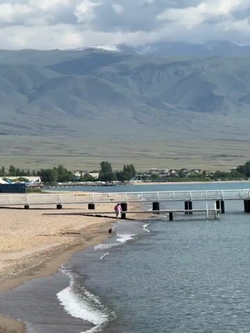 A person in a pink jacket stands on the shore of a lake or ocean, near a pier. The water is calm with gentle waves lapping at the sandy beach. The pier is white and extends out into the water, with several support structures visible. In the background, there are mountains, some of which are capped with snow. The sky is cloudy. The person appears to be looking at the water.