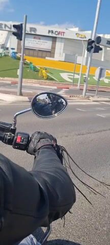 The video depicts a first-person perspective of someone riding a motorcycle on a road. The rider is wearing black leather gloves and a black leather jacket. The motorcycle has a chrome rearview mirror, which reflects the rider. The road is paved and marked with a red and white curb. There are cars on the road and palm trees lining the side. In the background, buildings and signs are visible. The weather appears to be sunny.