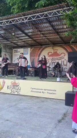 A band performs on an outdoor stage. On the left, a man plays a red electric guitar. Next to him, a man plays an accordion and sings into a microphone. To their right, another man plays a black electric guitar. Next to him, a woman with long dark hair, wearing a black dress with red embroidery, sings into a microphone. Behind the woman, a man plays the drums. The stage has a corrugated metal roof supported by metal beams. A yellow banner with text and a logo is draped across the front of the stage. A woman in a pink top with flowing sleeves stands in the foreground, facing the stage and extending her arms. A woman in a yellow top is partially visible on the far left. The ground is paved with concrete. Trees are visible in the background.
