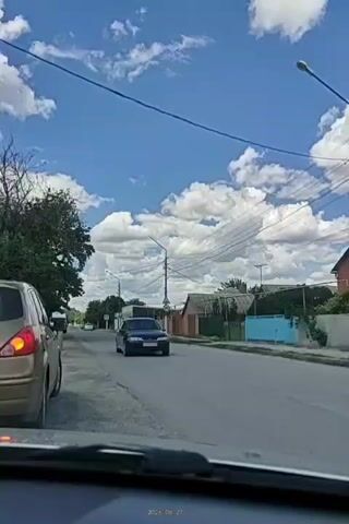 This media file captures a view from inside a vehicle driving down a street in Tatsinskiy, Russia. The scene is set on a clear, sunny day with scattered, fluffy white clouds in a vibrant blue sky. Trees line the left side of the road, and houses with visible roofs and fences are situated on the right. Several utility poles with electrical wires crisscross the sky.

The primary subject is the road and its surroundings. A gold-colored car is parked on the left side of the road, and a dark sedan is driving on the right side of the road, moving away from the camera. The activity is a car journey through a residential area.

Notable details include the bright, sunny weather, suggesting daytime. The presence of houses and utility poles indicates a populated, possibly rural or suburban, setting. The overall mood is calm and ordinary, reflecting a typical day.

Visible text includes a timestamp at the bottom of the frame: "2026.06.27".