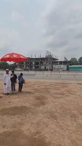 A group of five people stand next to a “National Food Village” sign. A red umbrella with a logo shades them. The sign indicates the restaurant is a highway restaurant in Dhopakandi, Raosidabad, Ullipara, Sirajganj. The sky is overcast. Behind the group, a highway carries trucks and a bus. Further back, a building is under construction. The ground is a light brown dirt. One person is looking at their phone, one is wearing a red dress, one is wearing white clothing, and two other individuals are also standing.