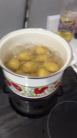 The video shows a pot of potatoes boiling on a stovetop. The pot is white with a floral pattern around the rim. Several small potatoes are submerged in bubbling water. In the background, a bottle of oil is visible. The overall environment appears to be a kitchen.