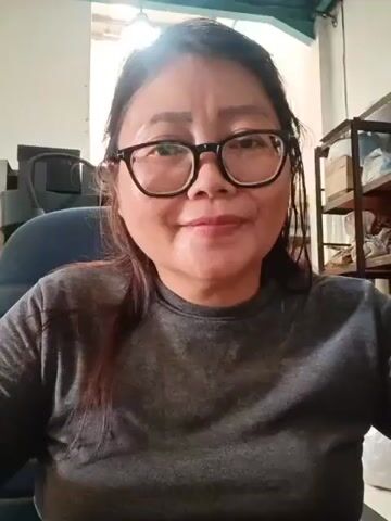 The video displays a medium close-up of a woman with dark hair, wearing glasses and a gray t-shirt. The lighting is bright, with a strong light source coming from the upper left, creating a lens flare effect. The woman's eyes are closed and she appears to be smiling or in a state of deep thought. She is seated in what appears to be an office or indoor setting, with a chair and shelving units visible in the background.
