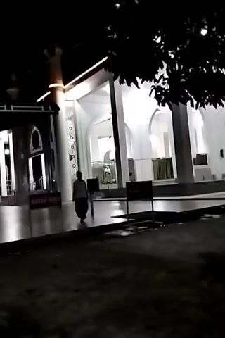 A short video clip captures a nighttime scene outside a white building with arched entrances and columns. The building appears to be a mosque, with a minaret visible on the left. Several people are present, dressed in traditional Islamic attire. One person in the foreground is wearing a white cap and a dark, long garment. Others are seen walking or standing in the background. A sign with red lettering is placed near the entrance. The ground is paved and wet in some areas, suggesting recent rain. Trees with dark foliage overhang the scene from the right, creating shadows. The lighting is artificial, casting bright illumination on the building and its surroundings. The video appears to be recorded from a slightly elevated position, looking down towards the building.