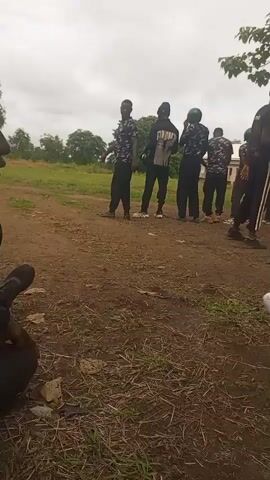 The video shows a group of young men, possibly cadets or trainees, standing in an outdoor area. They appear to be in uniform, with some wearing camouflage tops and black pants, while others are in black shirts with white stripes on the pants. One individual in the foreground is wearing a black shirt with "POLICE" written on the back. They are standing on a dirt path with dry grass and leaves on the ground. In the background, there is greenery, trees, and a cloudy sky. Some of the individuals are holding objects, possibly for training or equipment. The overall atmosphere seems to be one of instruction or preparation.