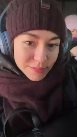 A woman is seated, possibly on public transport. She wears a burgundy beanie, a burgundy scarf, a dark jacket, and blue headphones. She glances down and then looks directly into the camera, maintaining a slight smile. The background is blurry and indistinct.