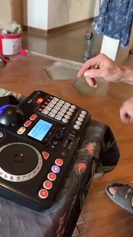 The video shows a person's hands interacting with a black DJ controller that is placed on a table. The controller has a circular platter on the left, a small screen in the middle, and numerous buttons and sliders. The person's fingers are seen pressing various buttons on the controller, suggesting they are operating the device. The environment appears to be indoors, with a wooden floor and a blurred background that suggests a room with some construction or renovation materials, including what looks like a bucket and a power tool. The overall lighting is moderate.