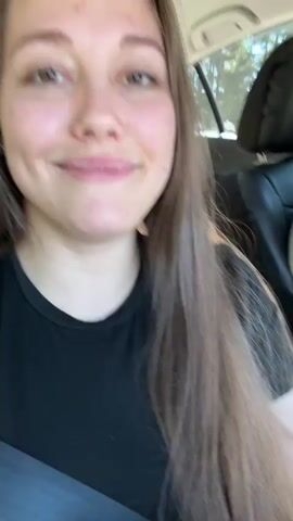 A woman with long brown hair and light skin is in the passenger seat of a car. She is smiling and winking at the camera, holding up two fingers in a peace sign. The man in the driver's seat has curly brown hair and is looking away from the camera. The car appears to be moving, as evidenced by the blurry background.