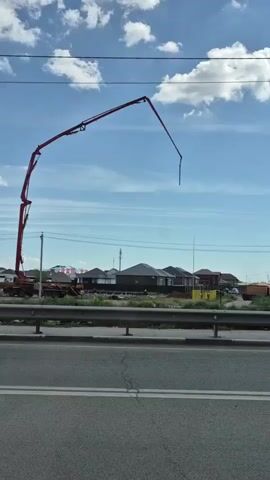 A concrete pump boom truck is positioned on the left side of the frame. The boom is extended upwards and to the right, reaching a significant height. The truck is situated in what appears to be a construction area, with several houses under construction visible in the background. The houses are mostly dark-colored with visible roofs. A metal barrier runs horizontally across the middle of the frame, separating the construction site from a road in the foreground. The road has two white lines painted on it. The sky is blue with scattered white clouds. A silver car is visible on the road in the bottom left of the frame, driving past. Overhead wires are present in the upper portion of the image.