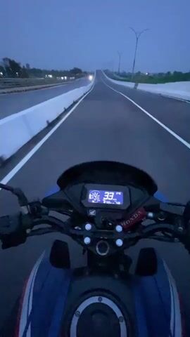The video captures a first-person view from a motorcycle, driving on a straight, empty road during dusk or early night in Rupganj, Bangladesh. The motorcycle's blue and black tank is visible in the foreground, along with its digital dashboard. The speedometer shows the speed increasing from 33 km/h to 51 km/h as the motorcycle accelerates. The "HONDA" brand name is visible on the handlebar. The road ahead is well-paved with clear white lane markings and white barriers on both sides. Streetlights are visible along the sides of the road, and distant lights can be seen in the background, suggesting either other vehicles or distant structures. The sky is a deep blue, indicating the transition from twilight to night.