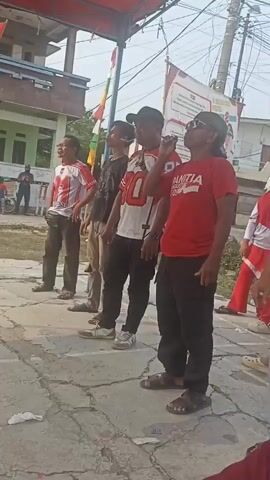 The video shows an outdoor event with several people, mostly men, standing in a line. They appear to be participating in some sort of competition or activity. The person in the foreground on the right is wearing a red t-shirt that says "PANITIA" which means "committee" in Indonesian. He is holding something up to his mouth, possibly a small object. The other men in the line are looking upwards. In the background, there are buildings, trees, and electrical wires. There are also some flags and banners, suggesting a festive or organized event. A woman wearing a black hijab and a white shirt is partially visible on the right side of the frame, facing away from the camera. The ground is paved with what appears to be concrete or asphalt.