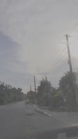 This is a first-person perspective video filmed from a vehicle, driving down a paved road lined with trees and utility poles. The sky is overcast, with the sun appearing as a bright, diffused orb, suggesting either early morning or late afternoon. The overall atmosphere is somewhat hazy and muted.

The scene depicts a rural or suburban road, likely in Merenii Noi, Moldova, given the location context. The road itself shows signs of wear, with visible patches and cracks. On either side of the road are trees and vegetation, with some properties hinted at by fences and structures in the background. The utility poles with numerous wires are a prominent feature, indicating the presence of infrastructure.

There are no people visible in the video, nor any specific activity other than the act of driving. The focus is on the environment and the road ahead. The weather appears to be cloudy, but the sunlight indicates it's daytime. There is no visible text in the video. The mood is neutral, conveying a sense of travel through a quiet, somewhat ordinary setting.