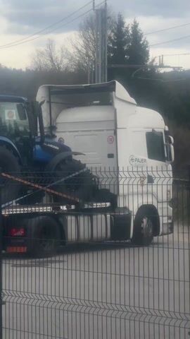A white Scania tractor truck is parked behind a chainlink fence. The truck is hauling a flatbed trailer, on which are loaded three blue Landini tractors. The tractors are secured to the trailer with straps. The truck is parked on a paved surface, and there are trees and power lines visible in the background. The sky is overcast. The truck cab has "Pajer" printed on the side.