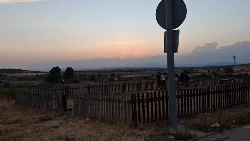 The video captures a wide shot of a rural landscape at dusk. In the foreground, a weathered wooden fence encloses a picnic area with a table and several people sitting and standing. The fence is made of vertical wooden planks with a rough, uneven finish. To the right of the fence, a metal pole with a circular sign is visible. Beyond the fence, the landscape stretches out with dry grass, scattered trees, and a dirt road. In the mid-ground, more trees and some buildings or structures can be seen. The background features rolling hills and mountains fading into the distance. The sky is filled with soft, pastel colors of sunset, with the sun partially obscured by clouds, casting rays of light through them. The overall atmosphere is peaceful and serene, indicative of an outdoor gathering during sunset.