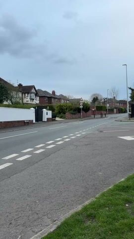 The scene is a street intersection on a cloudy day. On the left side of the street, there are houses with white walls and brick details. Across the street, houses are visible behind greenery and brick walls. A speed limit sign is posted near the intersection. The road has white dashed lines marking lanes and a large white arrow indicating direction at the intersection. A street lamp stands to the right. A patch of green grass borders the road on the right side of the frame.