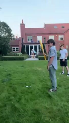 The scene shows four young men in a grassy backyard. One man, wearing a gray shirt and green pants, holds a yellow plastic bat. Another man, in a gray hooded sweatshirt and jeans, throws a small, white object toward him. A third man, wearing a white shirt and dark shorts, stands to the side. Behind them, a large red brick house with a sunroom is visible. The man with the bat swings and appears to hit the object. The person who threw the object jumps up and down, appearing excited. Litter is on the grass. The sky is a muted blue, suggesting it might be dusk.