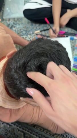 A close-up, overhead shot shows the back of a person's head with short, dark hair that swirls into a cowlick pattern. A hand with manicured nails points at the center of the cowlick. In the background, another person sits with legs crossed, drawing on a white sheet of paper with colored markers scattered around. Both are sitting on a patterned rug. The foreground person wears a light orange shirt.
