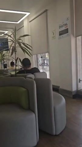 The camera pans slowly through a waiting area, where several people are sitting on round, light grey couches with green accents. The room is long and narrow with light-colored wood flooring, white pillars, and a high ceiling with square, recessed lights. There are several potted plants scattered throughout the area. On the right side of the room is a sign that says "Zona Digital".  Further down the hall, there are workstations with chairs and desk partitions. The camera pans from right to left, showing the arrangement of the seating, the people waiting, and the overall layout of the space.