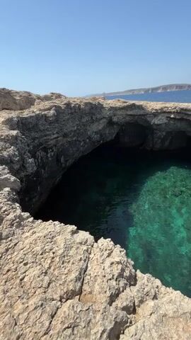 The video presents a breathtaking aerial-like view of a natural grotto or sea cave carved into rugged, light-colored rocky cliffs. The camera initially looks down into the grotto, revealing intensely clear, turquoise-green water that allows the rocky bottom to be seen distinctively. Darker shadows within the water suggest varying depths or areas obscured by the cave's overhangs.

As the camera slowly pans from left to right, it showcases the arch-like opening of the grotto, which leads out to the vast, deep blue expanse of the open sea. The foreground is dominated by the textured, dry rock surface of the cliff, with small patches of sparse green vegetation. In the mid-ground, the tranquil Mediterranean Sea stretches to the horizon under a perfectly clear, vibrant blue sky, indicating bright, sunny weather. A small white sailboat can be observed sailing in the distance on the open water. Another faint landmass is visible on the distant horizon, completing the coastal panorama.

The time of day appears to be mid-day or early afternoon due to the strong, direct sunlight. The scene depicts an untouched natural landscape, evoking a sense of serenity and natural beauty, consistent with the rugged coastlines of Mellieħa, Malta. No people or visible text are present in the video, though a brief, indiscernible male voice can be heard at the very start.