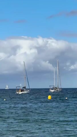 The video shows a serene ocean scene under a bright blue sky with scattered white clouds. Two sailboats are anchored in the calm, blue water, with their masts reaching towards the sky. A smaller sailboat is visible in the distance on the left. Yellow buoys are scattered in the water, marking the area. The waves are gentle, creating a peaceful atmosphere. The sun is shining, casting a bright light on the scene.