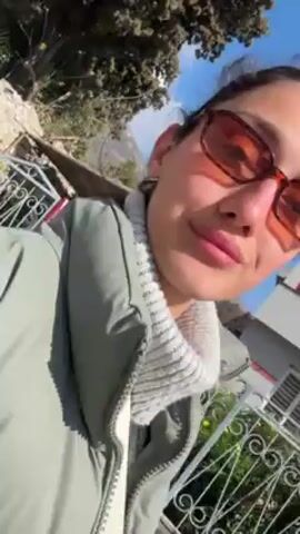 A woman is outdoors, facing forward and then turning to the side to face the right side of the frame. She has dark hair pulled back, wears dark-framed sunglasses, a white turtleneck sweater, a light green vest, and a white bag strap crossing her body. The background consists of green trees, a building with a red wall and white trim, a blue sky with clouds, and a white fence.  The video appears to be taken from a handheld device.
