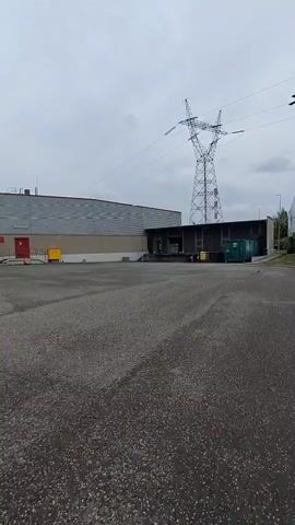 A handheld video begins with a low-angle shot of an asphalt lot in front of a long, gray industrial building with red doors and yellow bins. A tall, metal power line tower is visible in the background, along with a cloudy sky and some trees. The camera pans slowly to the left, revealing more of the building and a chain-link fence. It then pans right, showing a wooden fence and a residential building. The video concludes with a wider view of the lot and the power line tower.
