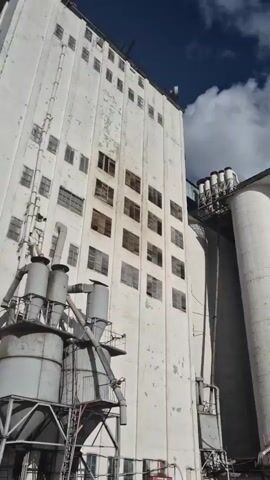 This video showcases an industrial complex, likely a grain elevator or processing facility, in Petropavl City Administration, Kazakhstan. The scene is dominated by large, cylindrical concrete silos painted white, with a tall, multi-story building adjacent to them. The architecture suggests an older industrial design, with numerous windows on the taller structure and various external pipes and machinery.

The activity appears to be a slow pan across the exterior of these industrial buildings. The camera movement is smooth, suggesting a deliberate filming of the location. The time of day is likely late morning or early afternoon, indicated by the bright sunlight and the shadows cast by the structures. The weather is partly cloudy, with large, dramatic white clouds filling a bright blue sky. Sunbeams are visible, breaking through the clouds in some shots, creating a sense of atmospheric depth.

There are no visible people in the immediate vicinity of the silos in the first few frames, but in the final frame, a lone individual is seen walking away from the camera in a large, open, paved area in front of the industrial buildings. This area appears to be an industrial yard, with some patches of dirt and debris. The overall mood of the video is somewhat somber and stark, reflecting the utilitarian nature of the industrial setting. There is no visible text in the video.