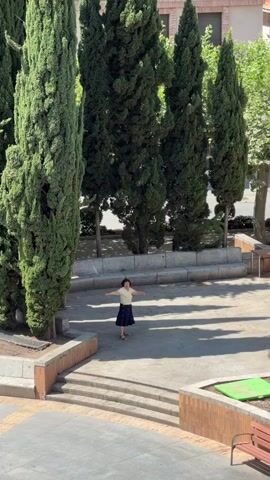 A woman in a yellow top and a dark blue skirt with a floral pattern stands in a paved outdoor area surrounded by tall green trees. She is facing the camera with her arms outstretched to her sides, then lowers them. She moves her arms as if dancing or gesturing. The area appears to be a public space with tiered seating in the background. Sunlight casts shadows on the ground.